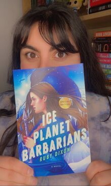 Antigoni Selena, holding up the book, Ice Planet Barbarians. The cover features a woman and blue horned alien character surrounded by snow.